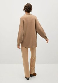 Maglione lavorato a maglia beige oversize con spalle abbassate, dettagli di bottoni sulle maniche, abbinato a pantaloni beige coordinati e scarpe marroni.