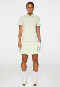 Ljusgrön pikétröja med korta ärmar, matchad med en veckad kjol. Modellen bär vita strumpor och golfhandske. Mjuk, slät textur.