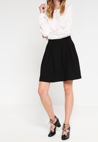 Blusa bianca con dettagli in pizzo abbinata a una gonna nera plissè, completata da scarpe col tacco nere con dettaglio allacciato.