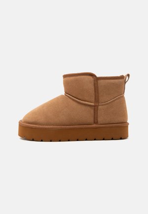 Brun ruskind ankle boot med tyk gummisål og syningsdetaljer på en ensartet lysegrå baggrund.