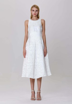 Femme blonde portant une robe midi sans manches blanche avec une texture florale et un col orné de bijoux, associée à des talons blancs à bride de cheville, se tenant immobile.
