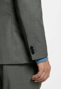 Grå, struktureret jakkesætjakke med en slank pasform, tre sorte knapper ved manchetten og et blåt strikket indvendigt lag, der er synligt ved ærmet.