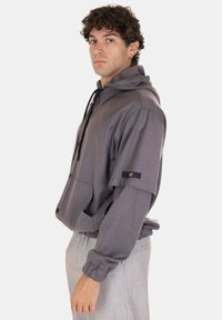 Giacca grigia con cappuccio, caratterizzata da un'apertura con bottoni, silhouette oversize, cordini regolabili e un accento con logo nero sulla manica.