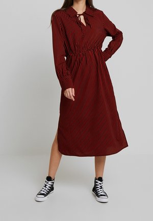 Vrouw die een rood-zwart gestreepte midi-jurk met lange mouwen, een keyhole-halslijn en zwarte hoge sneakers draagt.