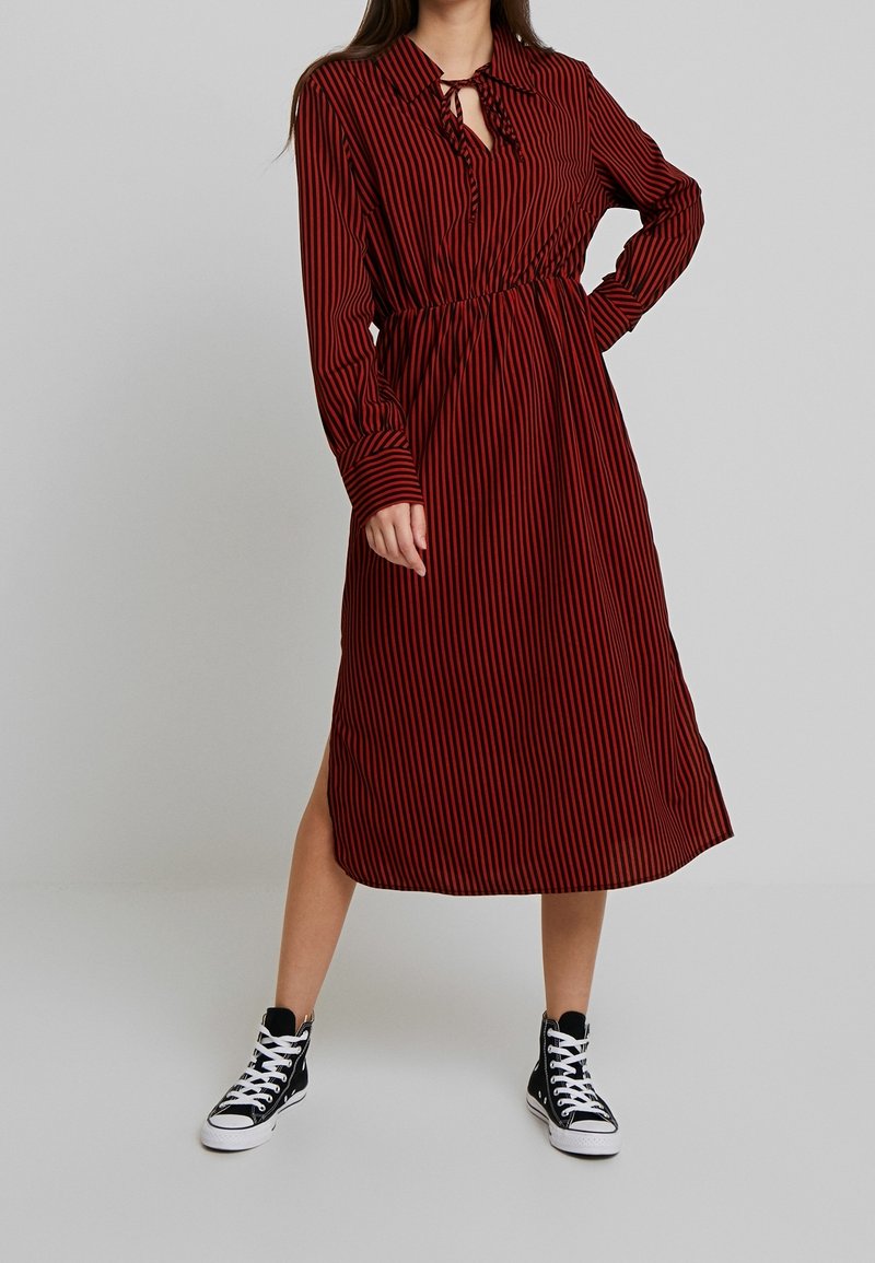 Femme portant une robe midi à manches longues rayée rouge et noire avec un col clé et des baskets montantes noires.