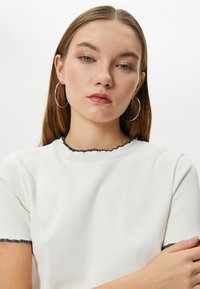 Hvid ribbet kortærmet top med sort flæse trim ved udskæringen og ærmerne. Model med langt hår og hoops øreringe. Enkel design.