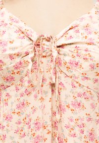 Rochie bej cu model floral în nuanțe de roz, portocaliu și albastru, având un detaliu cu șireturi împletite la piept cu panglici din material legate.