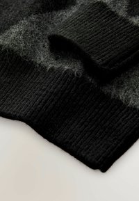 Maglia di lana nera e grigia con maniche e polsini dalla texture soffice su una superficie bianca.