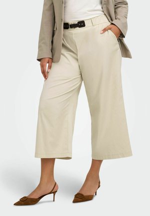 Frau trägt beige weite, gekürzte Hosen, braune spitze Slingback-Pumps, weißes Oberteil und beige Blazer mit Hand in der Tasche.