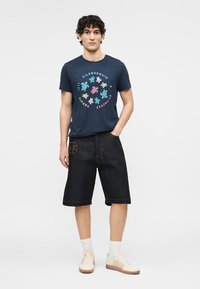 Joven de pie con la mano en el bolsillo, vistiendo una camiseta gráfica azul marino con tortugas, pantalones cortos largos negros, calcetas blancas y zapatillas beige.