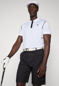 Biely polo tričko s čiernymi akcentmi, zipsovým golierom, čierne šortky a biely opasok; model drží golfovú palicu, na hlave má bielu čiapku a rukavicu.