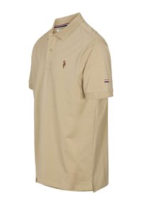 Smėlio spalvos polo marškinėliai iš medvilnės, turi trijų sagų užsegimą, siuvinėtą logotipą ir dryžuotas detalės ant rankovės. Lygus paviršius.