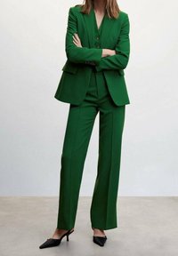 Green tailored suit with a fitted blazer and straight-leg trousers. Fabric appears smooth, with a structured design and three buttons on the blazer.