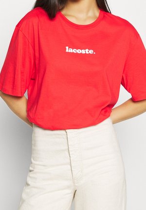 Czerwona bawełniana koszulka z krótkimi rękawami i okrągłym dekoltem, z białym logo "lacoste." umieszczonym na środku klatki piersiowej.