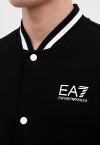 Κοντινό πλάνο μαύρου μπουφάν με λευκά κουμπιά και μαύρο-λευκό ριγέ γιακά που δείχνει το λογότυπο EA7 Emporio Armani στο στήθος.