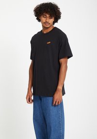 Zwarte katoenen t-shirt met korte mouwen, met een oranje logo op de borst. Gecombineerd met blauwe losse spijkerbroek.