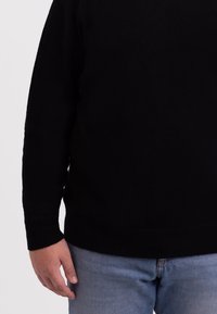 Maglione di maglia nera con scollo rotondo e polsini a coste, indossato sopra jeans azzurri chiari. La texture è morbida e uniforme.