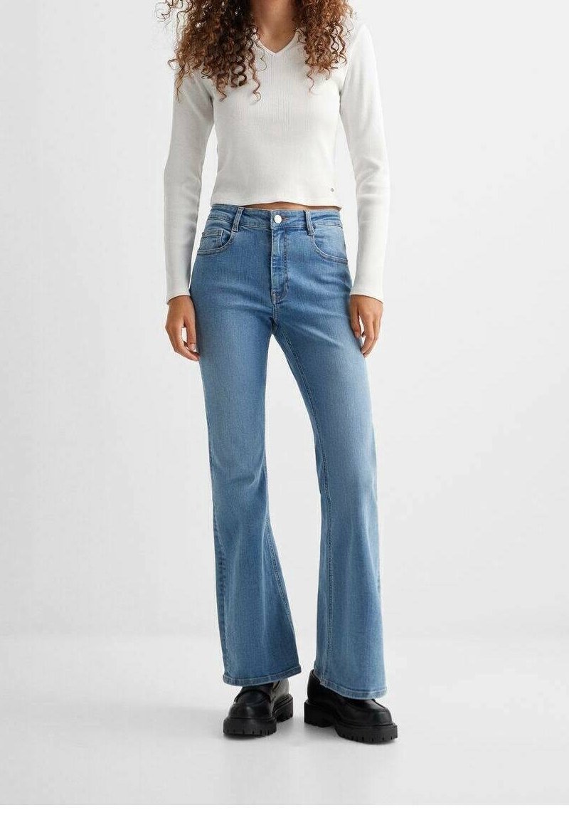 Femme portant un jean flare taille haute bleu clair, un crop top blanc à manches longues et des bottes noires épaisses, debout devant un fond uni.