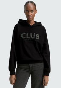 Schwarzer Hoodie mit strukturiertem silbernen "CLUB"-Text, aus Baumwollmischung, mit einer vorderen Kängurutasche und gerippten Bündchen. Kombiniert mit dunkelgrauen Jeans.