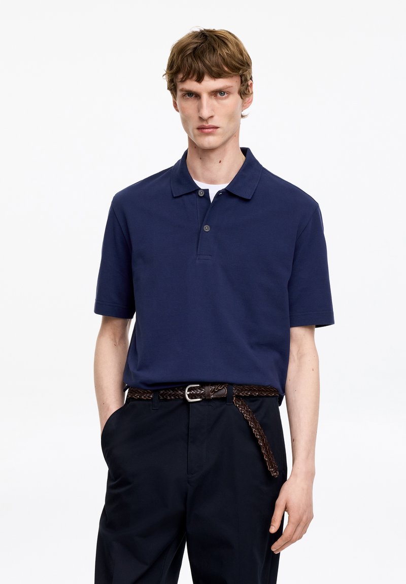 Jeune homme portant un polo bleu marine à manches courtes, un pantalon noir et une ceinture tressée marron, debout avec une main dans la poche.