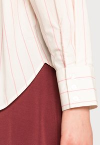 Chemise blanche à rayures verticales rouges, avec col replié et poignet boutonné. En dessous, un tissu bordeaux suggère un style superposé.