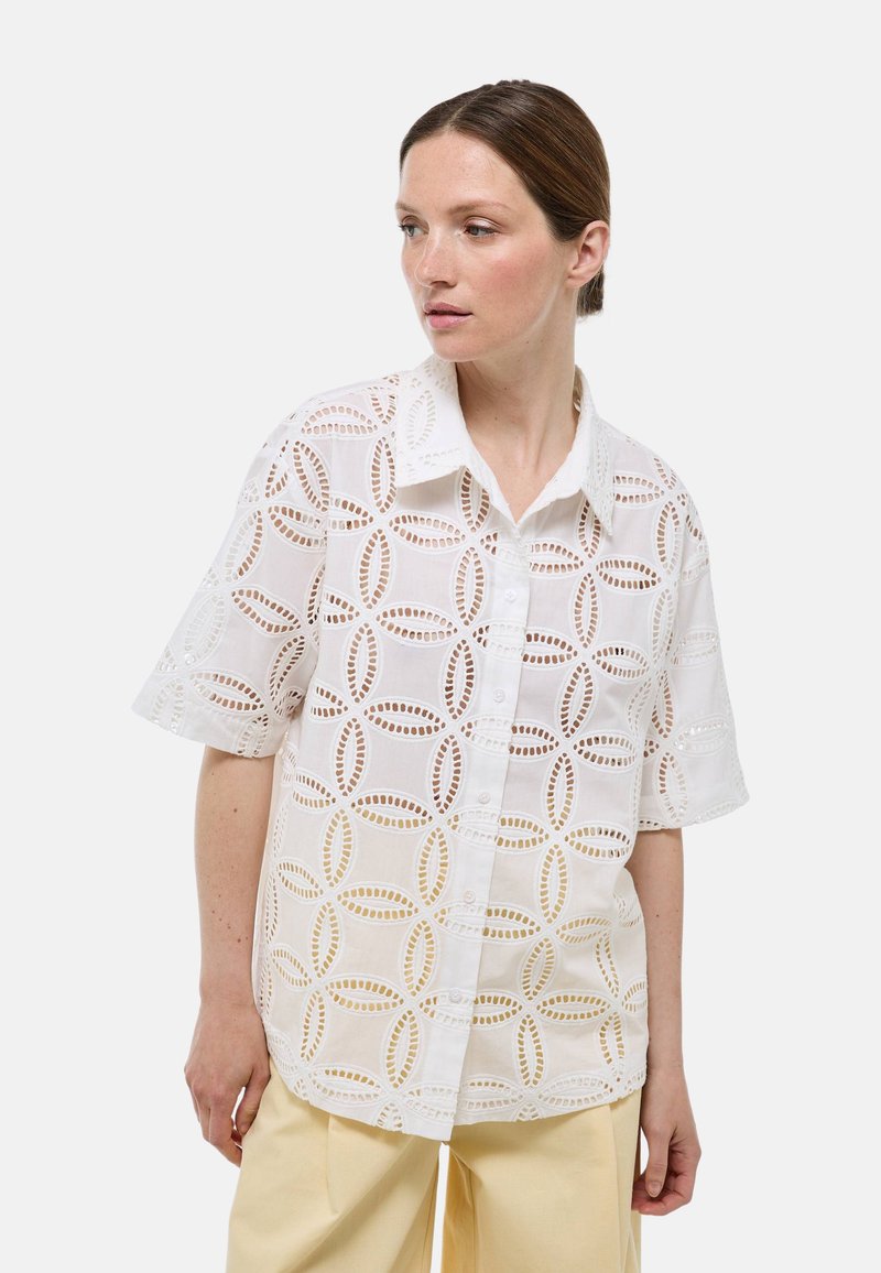 Chemise blanche à manches courtes en tissu léger avec des broderies et des motifs de découpe circulaires, dotée d'un devant à boutons.