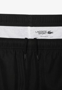 Svarta sportshorts med elastisk midja, dragsko och nättyg. Varumärkesetikett "LACOSTE SPORT" synlig på insidan.
