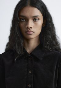 Schwarzes Cordhemd mit klassischem Kragen, Knopfverschluss und sichtbarem Strukturstoff. Schlichtes Design vor natürlichem Licht.