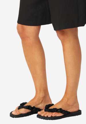 Piernas con pantalones cortos negros hasta la rodilla y sandalias planas negras con textura y tiras fruncidas sobre un fondo blanco.