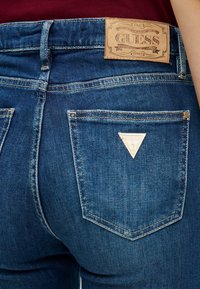 Plave traperice s teksturiranim završetkom, s tamnosmešnim kožnim oznakom s natpisom "GUESS" na stražnjem dijelu struka i trokutastim logotipom na džepu.
