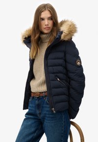Navy puffer jacket with faux fur hood, quilted design, and zippered side pocket. Paired with a beige knit sweater and blue denim jeans.