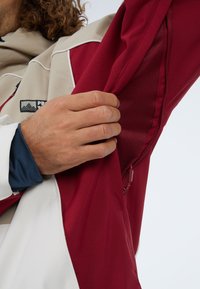 Rode en beige jas met gemêleerde voering. Voorzien van een ritszak onder de arm, mesh details en een logo patch op de borst.