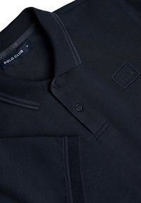 Polo de color azul marino hecho de tejido texturizado con cuello y dos botones. Presenta un logo cuadrado de "POLO CLUB" en la parte superior izquierda.