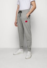 Pantalones de chándal grises de suave algodón, con cintura ajustable mediante cordón, bolsillos laterales y un parche de logo rojo en el muslo superior. Puños acanalados.