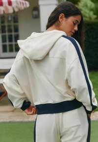 Krémová mikina s kapucňou, tmavomodré bočné pruhy, textúrovaná látka, voľný strih a rebrované manžety. Má klokánkovú kapsu a švy na chrbte.