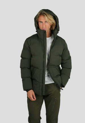 Homme portant une doudoune à capuche verte sur une chemise blanche et un pantalon cargo vert, debout devant un fond gris clair uni.