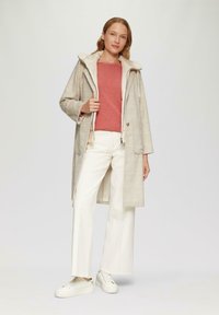 Beige geruite lange jas met een zachte crèmekleurige kraag, gecombineerd met een rode top en witte wijde broek. Witte sneakers maken de outfit af.