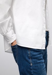 Hvid skjorte med pufærmer, parret med blå denimjeans med slim fit og synlige stingdetaljer.