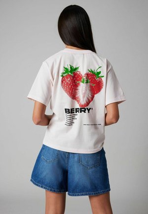 La camiseta de manga corta rosa presenta un gran gráfico de fresa y la palabra "BERRY". Combinada con pantalones cortos de mezclilla azules, que muestran detalles de costura visibles.
