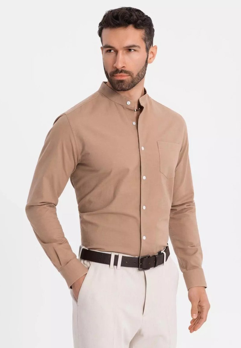 Chemise marron clair à manches longues avec un col montant, une poche poitrine unique et des boutons blancs. Tissu texturé associé à un pantalon de couleur claire.