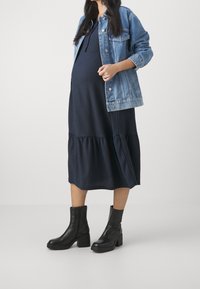 Ciężarna osoba w granatowej sukience z wieloma warstwami, nosząca dżinsową kurtkę i czarne botki za kostkę, podkreślająca warstwowy materiał i swobodny styl.