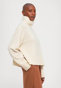 Łysy mężczyzna w kremowym swetrze z prążkowanym kołnierzem i brązowych spodniach, stojący bokiem z jedną ręką w kieszeni na białym tle.