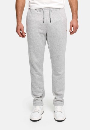 Mężczyzna w jasnoszarych spodniach dresowych ze ściągaczem w pasie i białych sneakersach, stojący na jednolitym tle.