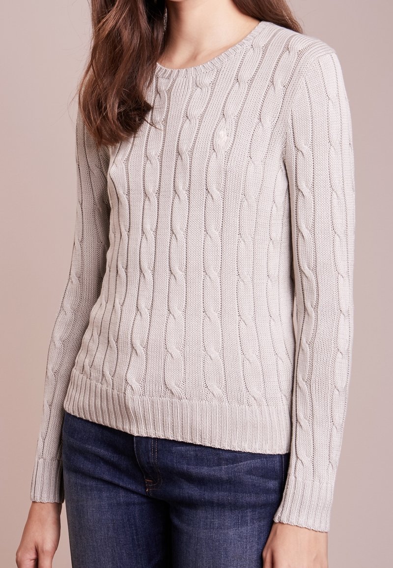 Femme portant un pull en maille torsadée gris clair et un jean bleu foncé, debout devant un fond beige uni.