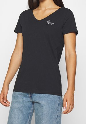 Femme portant un T-shirt noir à col en V avec le logo "Tommy Jeans" sur la poitrine et un jean en denim bleu clair, debout les bras détendus.
