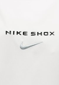 Μαύρο κείμενο NIKE SHOX πάνω από ένα μικρό ασημένιο κεντημένο λογότυπο Nike σε λευκό ύφασμα, πιθανότατα μέρος ενός ρούχου.