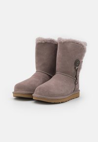 Botas de ante taupe de altura media con forro de piel suave, suelas de goma marrón y botón decorativo con charms colgantes en el lado exterior.