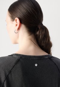 Top in tessuto nero con scollatura rotonda, caratterizzato da dettagli di cucitura visibili e un piccolo etichetta sul retro. I capelli sono raccolti in una coda di cavallo.