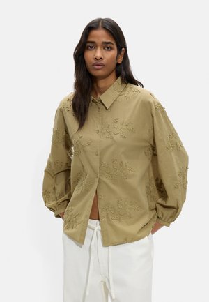 Kobieta o długich ciemnych włosach, ubrana w luźną, beżową koszulę z kwiatowym haftem i białe spodnie ze sznurkiem, stojąca na jednolitym tle.