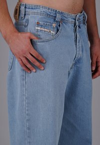 Hellblaue Denim-Jeans mit geradem Schnitt, kupferfarbene Beschläge und einem braunen Lederetikett am Bund. Die Taschen sind mit Nähten verziert.